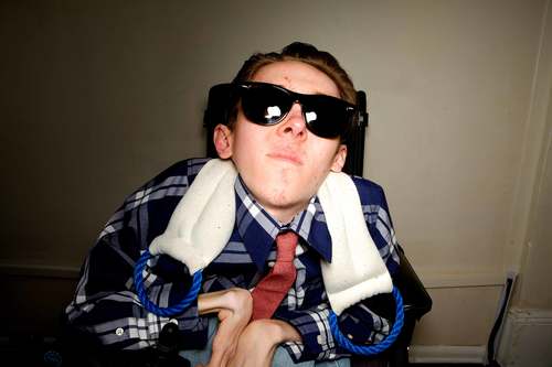 A young male with light skin using a wheelchair and wearing a blue shirt, a red tie, and sunglasses. Behind him, there is a tan wall. 