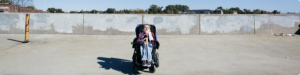 A young male with light skin using a wheelchair and wearing a blue shirt, a red tie, and sunglasses. Behind him is a tan wall.