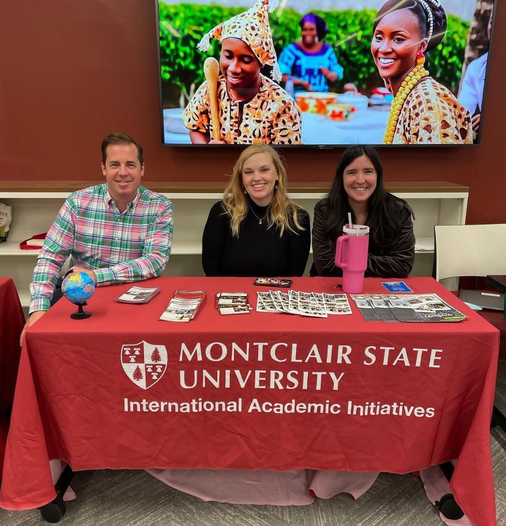 Group of three members of the Office of International Academic Initiatives in front of television with image of Burkina Faso cultural clothing. There is a man on the left side and two women to his right sitting in front of a table with a red tablecloth that has the office logo.