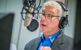 David Brancaccio wearing headphones speaking into microphone at Montclair State Studios.