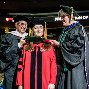 Graduate school student at commencement standing on stage being honored by dean and professor.