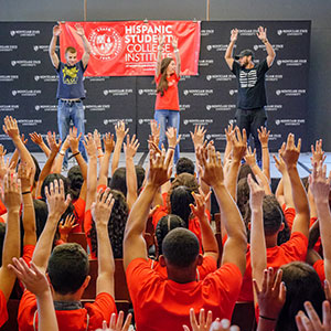 Students of Hispanic Student College Institute raising hands in air with performers on stage doing the same in front of them.