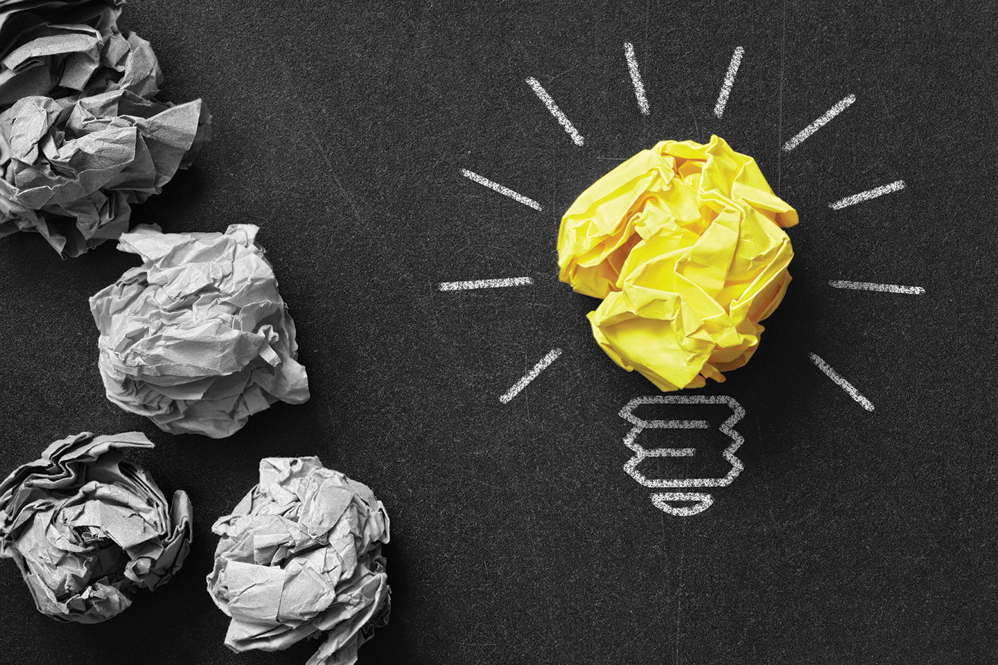 Blackboard with crumpled balls of paper, one of which is yellow and depicts a lightbulb.