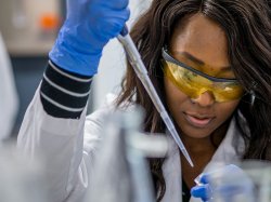 Student wearing lab coat and safety goggles injecting liquid into small container at the College of Science and Mathematics