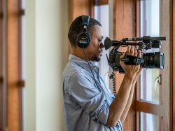 Student from School of Communication and Media wear headphones and holding a video camera filming through a window.