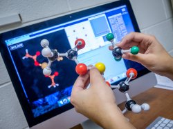 Hands holding models of chemical bonds in front of a computer display screen.