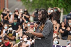 Jason Williams holding a microphone and speaking at an outdoor demonstration. a crowd is blurred in the background