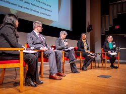 "Five people sit in chairs on a stage. One is speaking and holding a microphone."