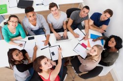 a group is seated around a table with paper and laptops working together. camera is above and they are turning their heads up towards the camera.
