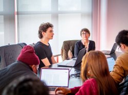 "Students sit around a table while a professor looks on in the background."