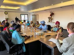 a large group of people sit around a conference table and listen and write as person at head of table speaks and gestures with hand