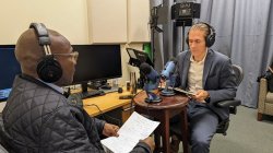 Two men have a conversation for podcast with microphones on the table. Both are wearing headphones and holding notes