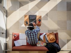 aerial photo of student sitting on couch, wearing headphones and working on computer
