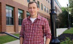 photo of professor Nicholas Williams in plaid shirt standing outside of a brick building