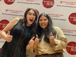Two smiling students giving a "thumbs up" in front of a backdrop with logos.