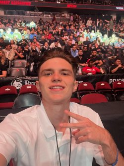 A student in a white t-shirt at a professional fighting event. He is smiling and happy and has a crowd behind him as the match gets ready to start.