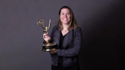 A woman dressed professionally, standing and holding an Emmy trophy against a dark backdrop.