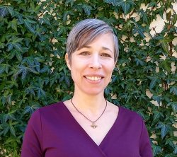 Christa Akers in purple shirt stands with greenery behind her