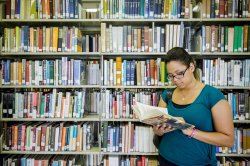 Student reading a book next to a shelf of books in the library
