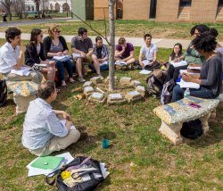 Photo of a classroom in an outdoor setting with faculty and students sitting on benches and the ground.