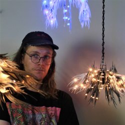 A white man wearing a navy blue baseball hat and a black shirt with a colorful graphic design stands behind lit, hanging pieces of art against a white background.