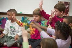 Image of children sitting on the floor in a classroom, raising their arms.