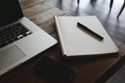 This is a picture of a macbook laptop, notebook, pen, and cellphone sitting on a wooden table