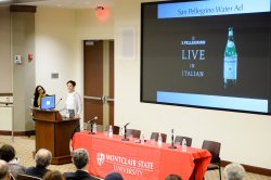 Photo of a presenter speaking in front of a projection screen.