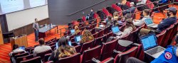 auditorium with white male faculty at front wearing facemask teaching to large group of students