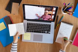 overhead photo of white female hands taking notes watching a video on a laptop lesson given by a white female teacher