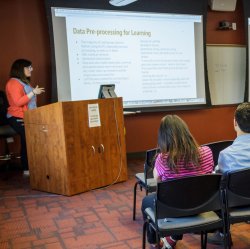 Image of a student in front of a podium presenting in front of an audience with a screen about computational linguistics in the background.