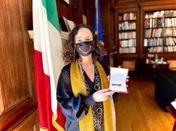 photo of Teresa Fiore with black face mask with white smile. Italian flag is in the background in an office with bookshelves