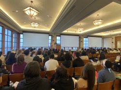 Full conference Center from back of room looking at stage with three projection screens and speaker standing at podium