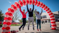Three individuals jumping underneath red and white balloon arch