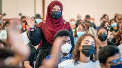 Woman in head wrap and face mask standing, holding up one hand