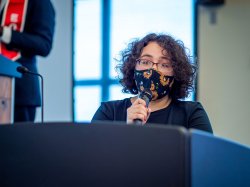 woman in floral-print face mask speaking into microphone behind podium