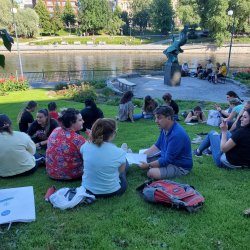 people sitting and talking on shaded lawn along a river