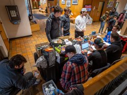 Male students sit and stand at a table stacked with canned goods