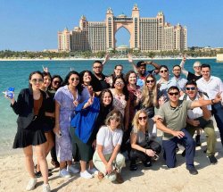 Group of students pose with water and large hotel in background.