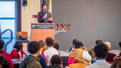 A man gestures with a microphone as dozens of students listen.