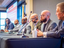 Six men sit at a table behind microphones.