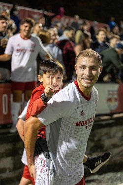 A man in a Montclair soccer uniform carries a child piggyback-style on the field.
