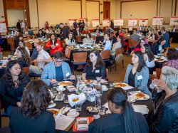 People seated at round tables eating lunch listen to a speaker during a symposium.