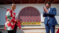 A woman wearing a white jacket with red flowers on it, holds an oversized pair of scissors and smiles after cutting a red ribbon as a man wearing a blue suit, smiles and applauds.