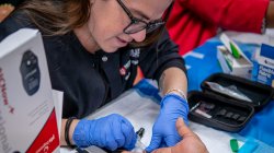 A woman with blue surgical gloves takes a fingerstick blood sample from a person’s finger.