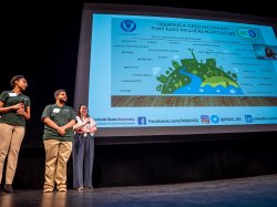 Three people stand in front of a large screen that has information about a green economy and agriculture on it.