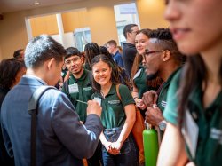Students in green shirts smile and talk with a man in a suit.
