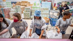 Students pack bags with food at a food pantry filled with pallets of canned goods.
