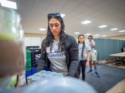A female student lifts a box of canned goods as other students line up behind her.