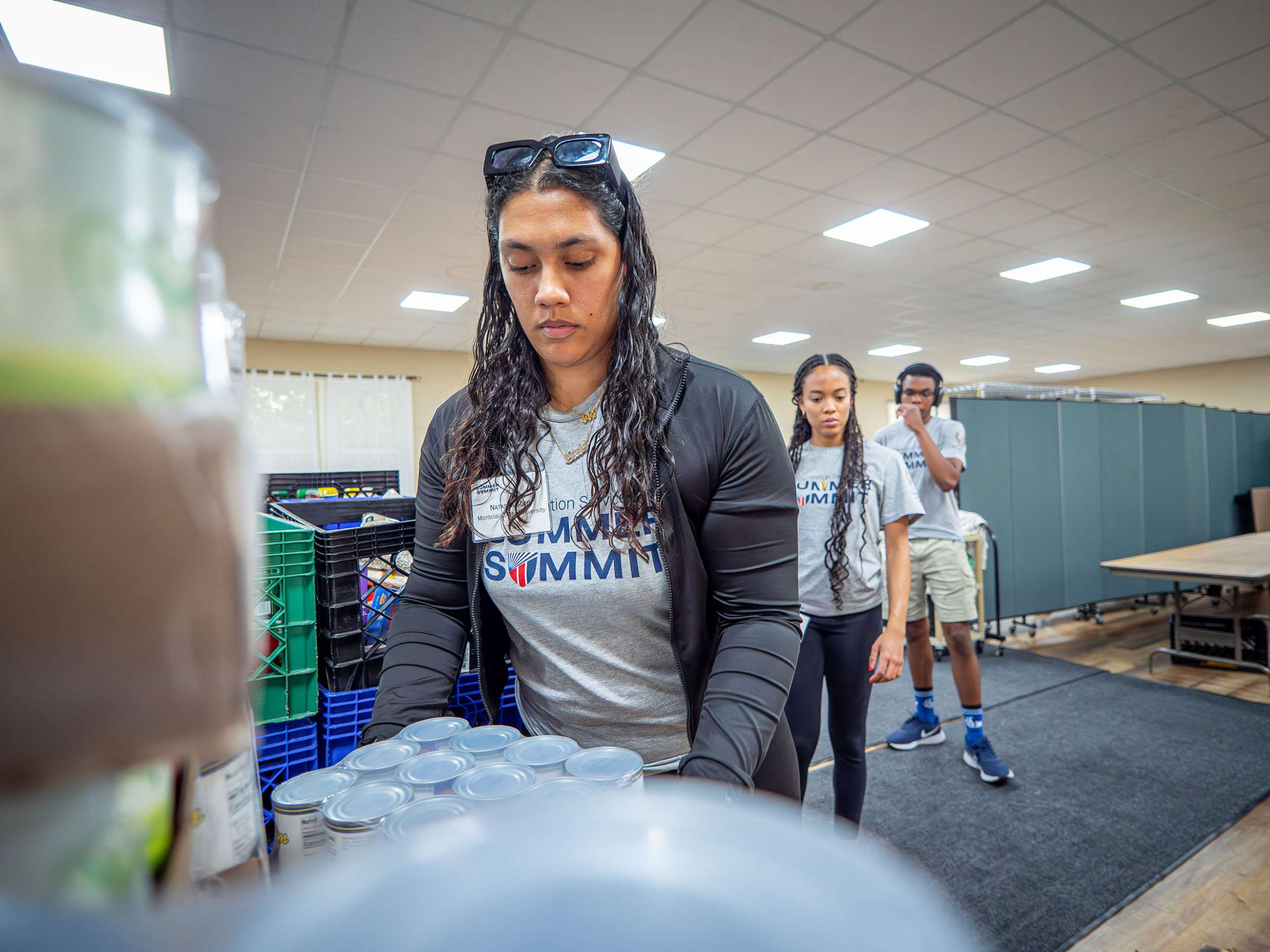 A female student lifts a box of canned goods as other students line up behind her.
